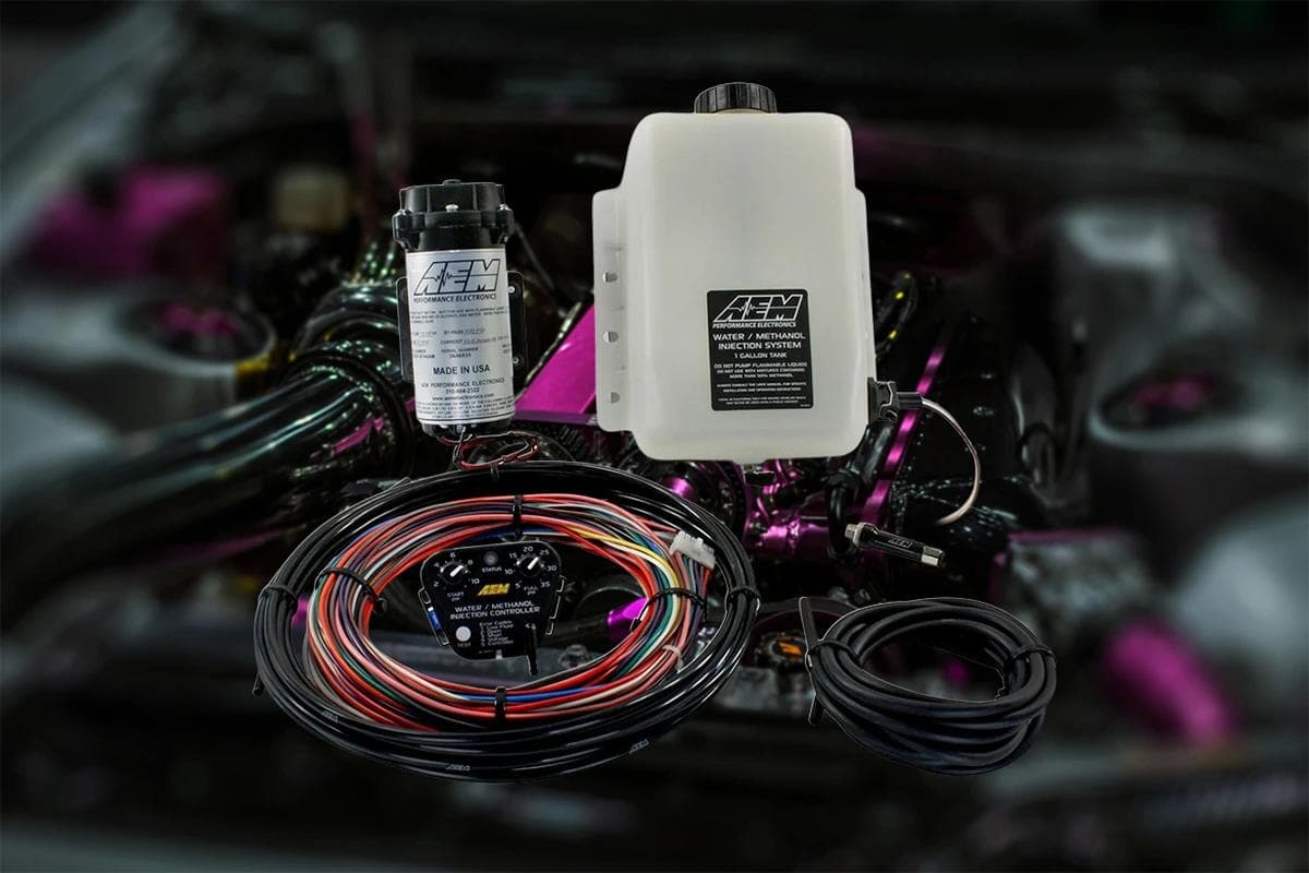 AEM water methanol injection kit components shown in an engine bay, including reservoir tank, pump, controller, wiring loom, and hoses.